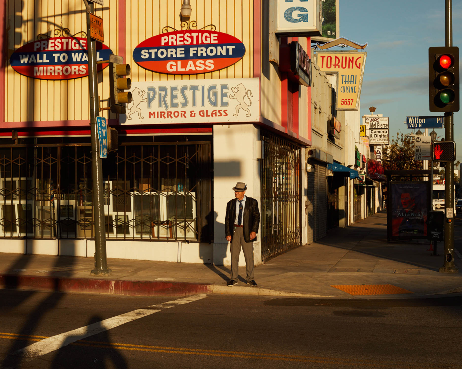 An adult male in a suit and hat stands on a city street sidewalk next to a mirror and glass store on a sunny day in a medium shot.