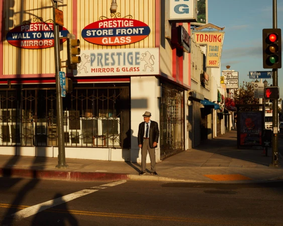 An adult male in a suit and hat stands on a city street sidewalk next to a mirror and glass store on a sunny day in a medium shot.
