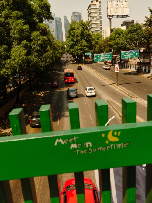 Traffic scene in Mexico City featuring a bus, cars on the road, and surrounding buildings viewed from a bridge with graffiti.