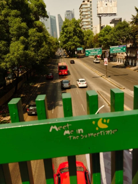 Traffic scene in Mexico City featuring a bus, cars on the road, and surrounding buildings viewed from a bridge with graffiti.