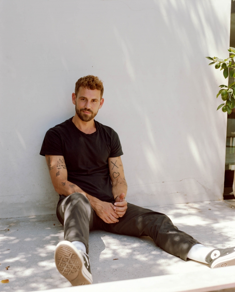 A young adult male with red hair and a beard poses while sitting on a floor in a black T-shirt and dark pants, lit by natural light.