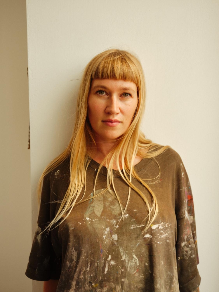 Portrait of a blonde adult woman with bangs, wearing an paint-splattered shirt, standing indoors against a white wall in natural lighting.