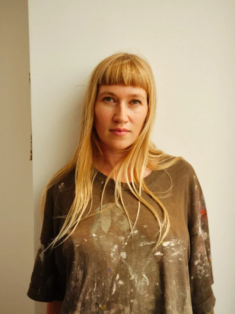 Portrait of a blonde adult woman with bangs, wearing an paint-splattered shirt, standing indoors against a white wall in natural lighting.