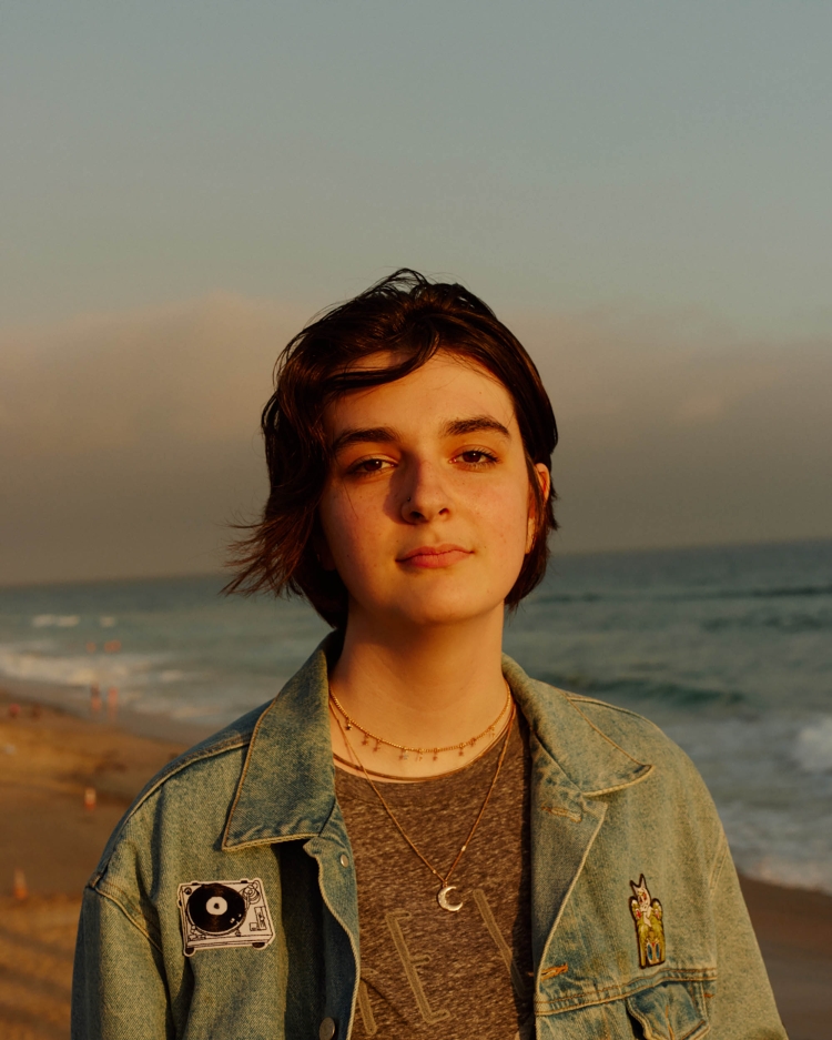 Teenage girl with short brown hair stands on a beach at sunset wearing a denim jacket, a gray t-shirt, and layered necklaces.