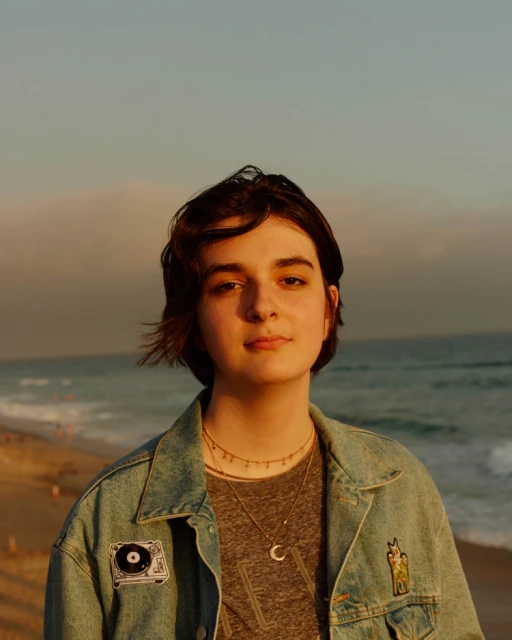 Teenage girl with short brown hair stands on a beach at sunset wearing a denim jacket, a gray t-shirt, and layered necklaces.