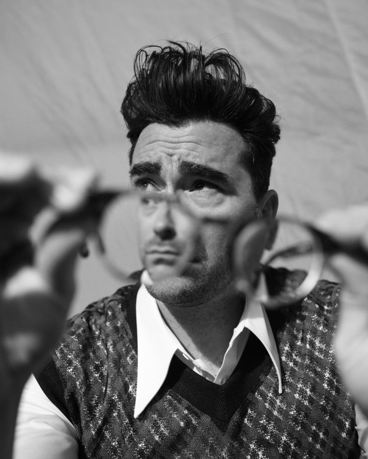 Black and white portrait of a man with dark hair holding eyeglasses, wearing a patterned vest and collared shirt against a light backdrop.