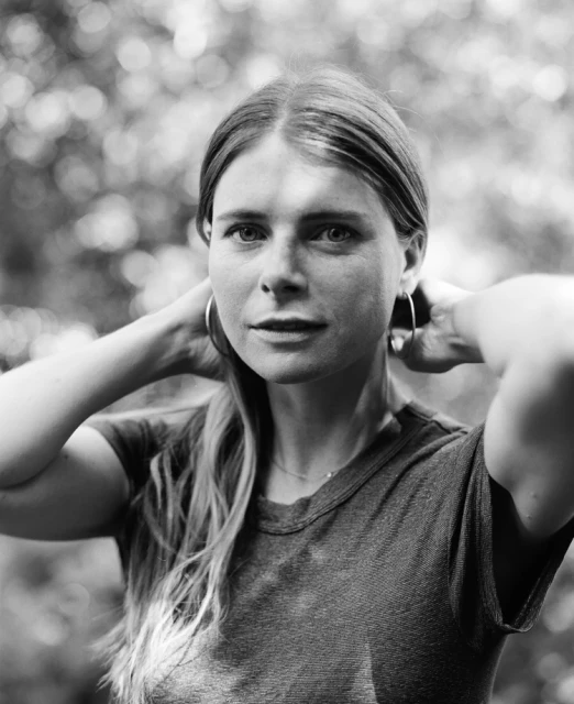 Black and white portrait of a young adult female with her hands behind her neck outside with a blurred foliage background.