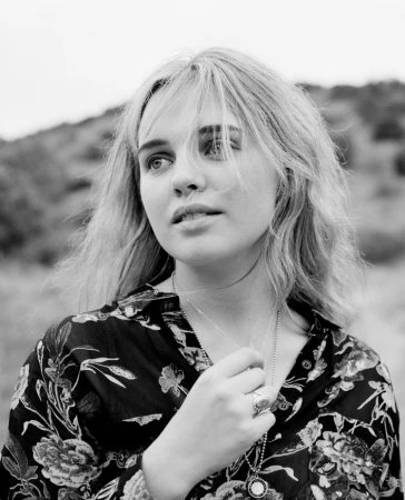 Portrait of a young blonde woman with a patterned dark shirt and necklaces against an outdoor natural landscape background.