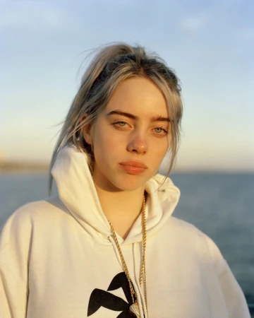 Portrait of Billie Eilish wearing a white hoodie and gold chain against an ocean backdrop at sunset