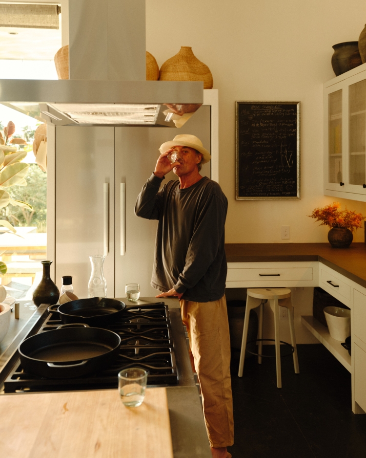 Man drinks from a glass in a kitchen with stainless steel appliances, wooden countertops, and woven basket decorations.