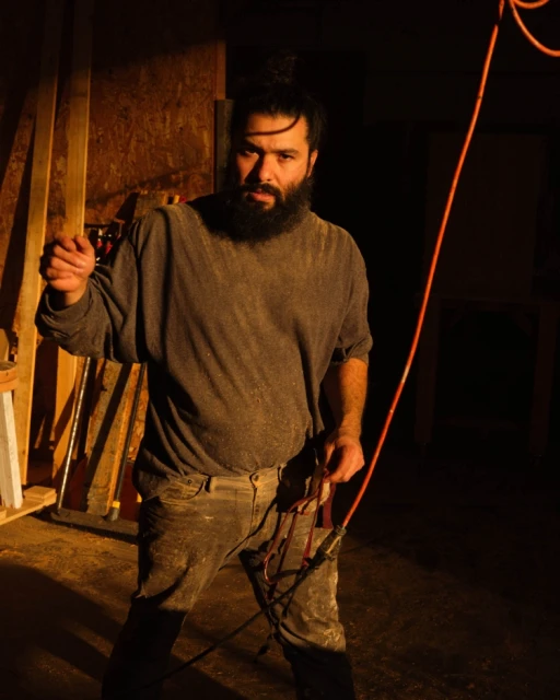 Bearded adult male stands indoors holding a coiled orange cord, showcasing rough, dusty clothing in a dimly lit interior space.