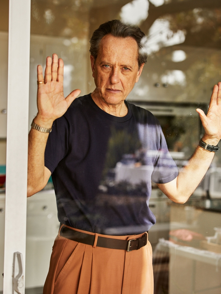 Portrait of a man standing behind a glass door, wearing a navy t-shirt and tan pants. His hands are up against the glass.