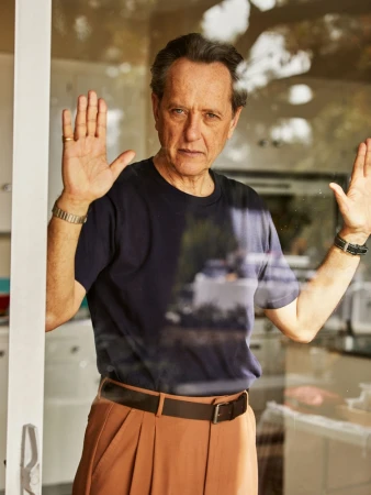 Portrait of a man standing behind a glass door, wearing a navy t-shirt and tan pants. His hands are up against the glass.