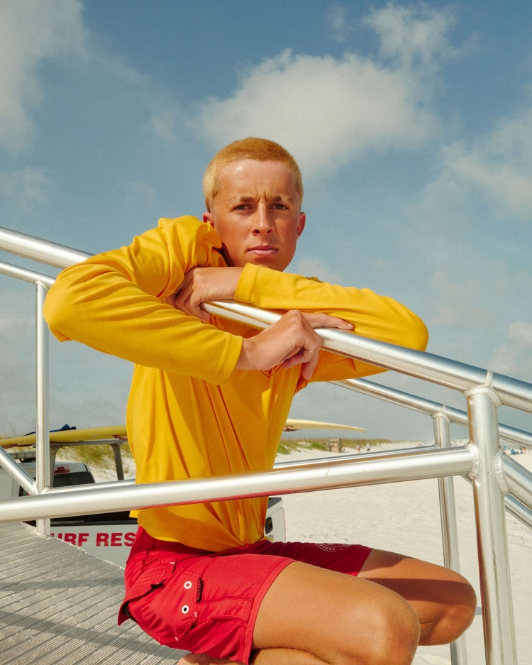 A lifeguard in a yellow shirt and red shorts poses on a beach access staircase, with sand, surfboards, and a blue sky in the background.