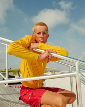 A lifeguard in a yellow shirt and red shorts poses on a beach access staircase, with sand, surfboards, and a blue sky in the background.