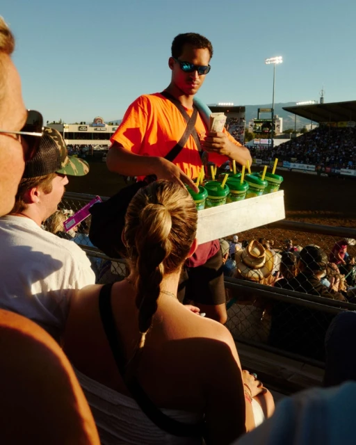 Man selling drinks at an outdoor stadium, wearing sunglasses and an orange shirt, with spectators and stadium seats in the background.