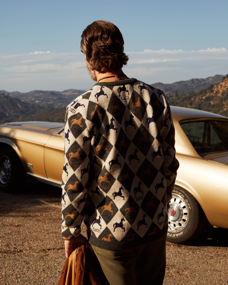 Adult male wearing patterned sweater stands near vintage gold Mustang on outdoor road, mountain landscape backdrop under blue sky.