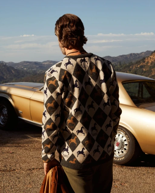 Adult male wearing patterned sweater stands near vintage gold Mustang on outdoor road, mountain landscape backdrop under blue sky.