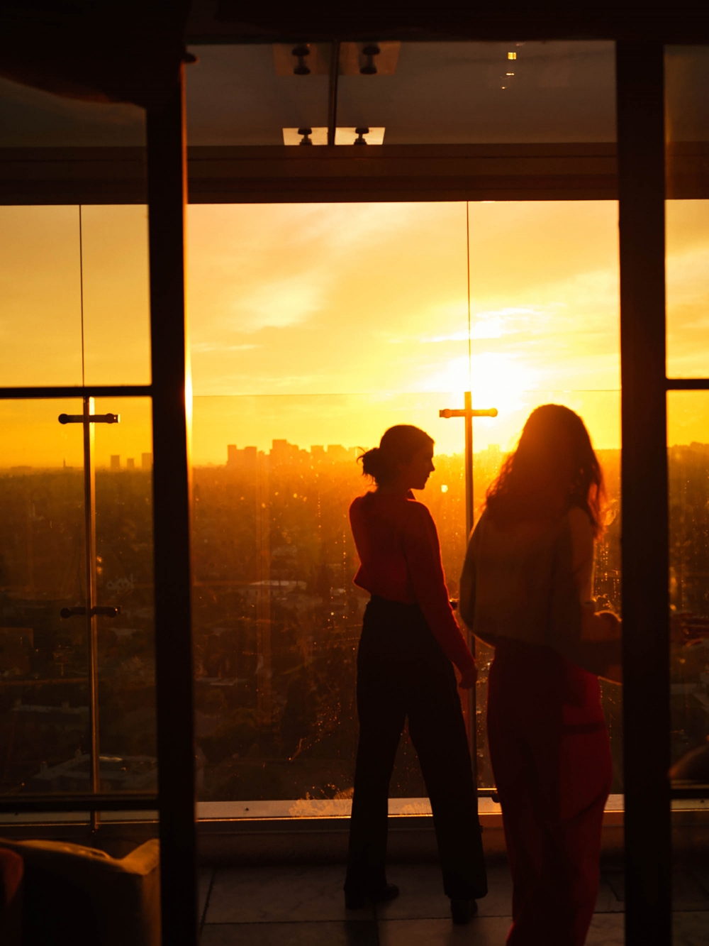Two women stand silhouetted in front of a city skyline at sunset seen through an indoor window frame with orange and gold sky.