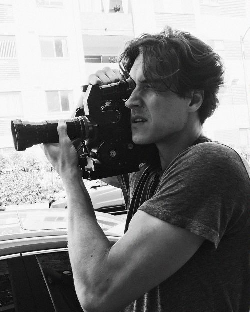 Monochrome shot of a dark-haired adult man holding a vintage film camera, wearing a gray t-shirt, with a building in the background.
