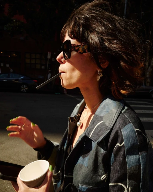 Woman with brown hair and sunglasses sips from a cup through a straw on a sunny day, wearing a patterned jacket outdoors.