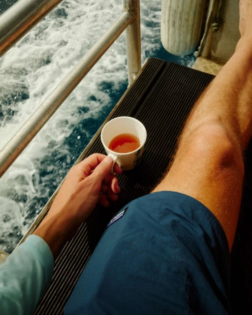 Man in blue shorts holds a cup of tea on a boat with foamy water and metal railing visible in the background.