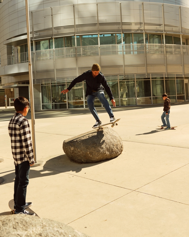 Skateboarder performs trick on rock in urban setting, observed by child in plaid shirt, another skateboarder in background.