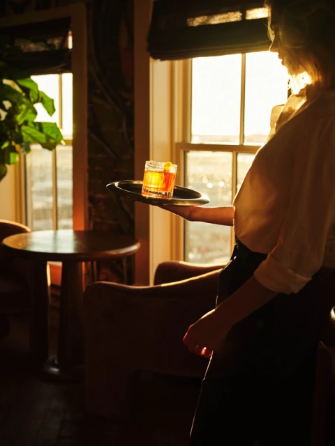 Bartender serves orange cocktail on silver tray in bar with warm light, wood furniture, and sunset window view. Drink photography.