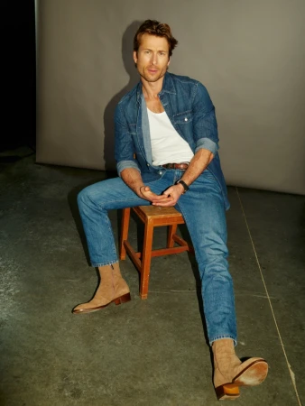 An adult male in a denim shirt and jeans, sitting on a wooden stool in a photo studio, looking at the camera with a neutral expression.