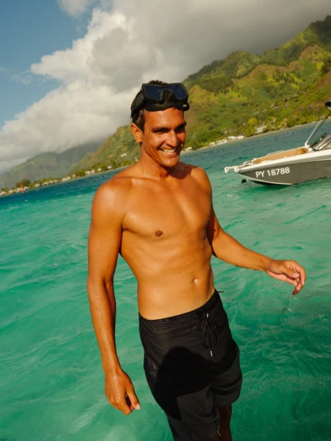 Smiling adult male in black swim trunks and goggles stands in clear turquoise ocean water with a motorboat and tropical landscape backdrop.
