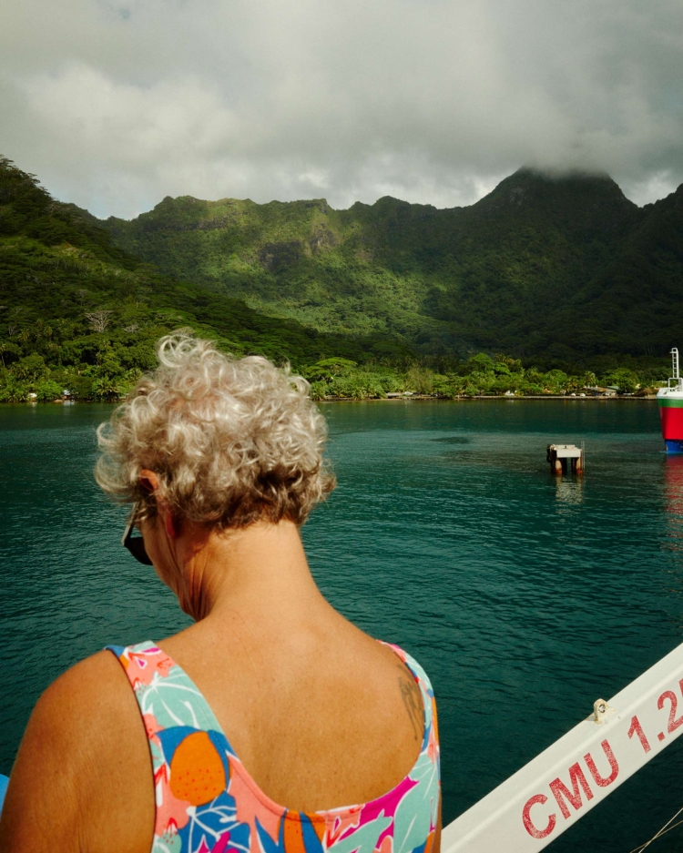 Back view of a woman in a floral swimsuit enjoying a tropical island view with green, mountainous landscape and calm blue water.