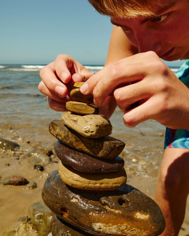 Young man balances stones on a sandy beach under a clear blue sky. Hands carefully place a rock atop a tower of brown stones.