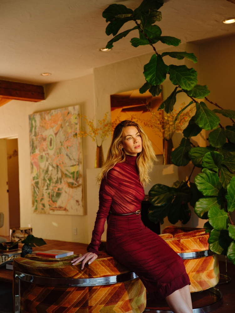 A blonde woman in a red dress sits on a patterned ottoman in a brightly lit room with artwork and greenery, creating an elegant portrait.