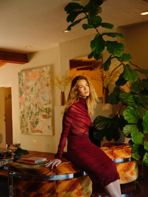 A blonde woman in a red dress sits on a patterned ottoman in a brightly lit room with artwork and greenery, creating an elegant portrait.