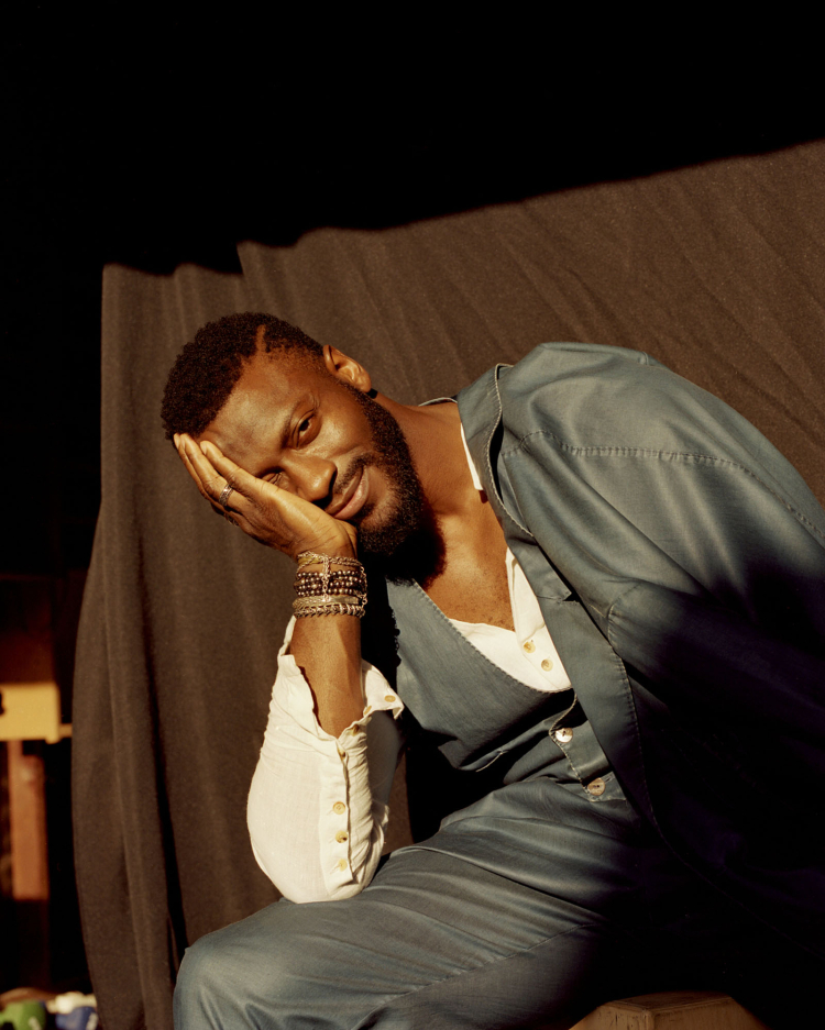 Portrait of a dark-skinned male model with a beard, wearing a jacket and bracelets, posing in a dimly lit photo studio against a draped backdrop.