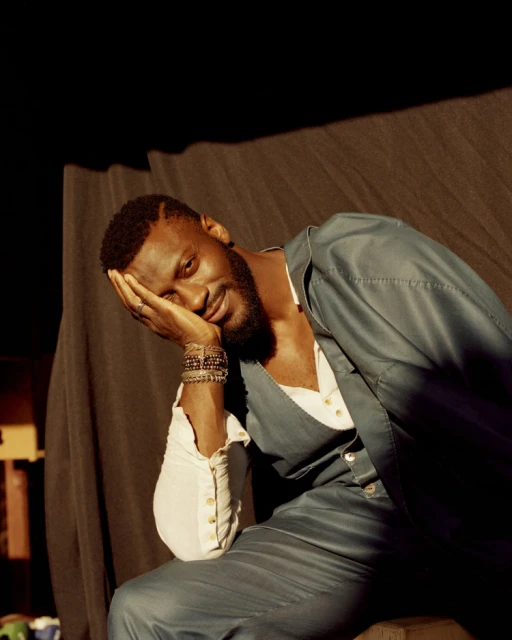 Portrait of a dark-skinned male model with a beard, wearing a jacket and bracelets, posing in a dimly lit photo studio against a draped backdrop.