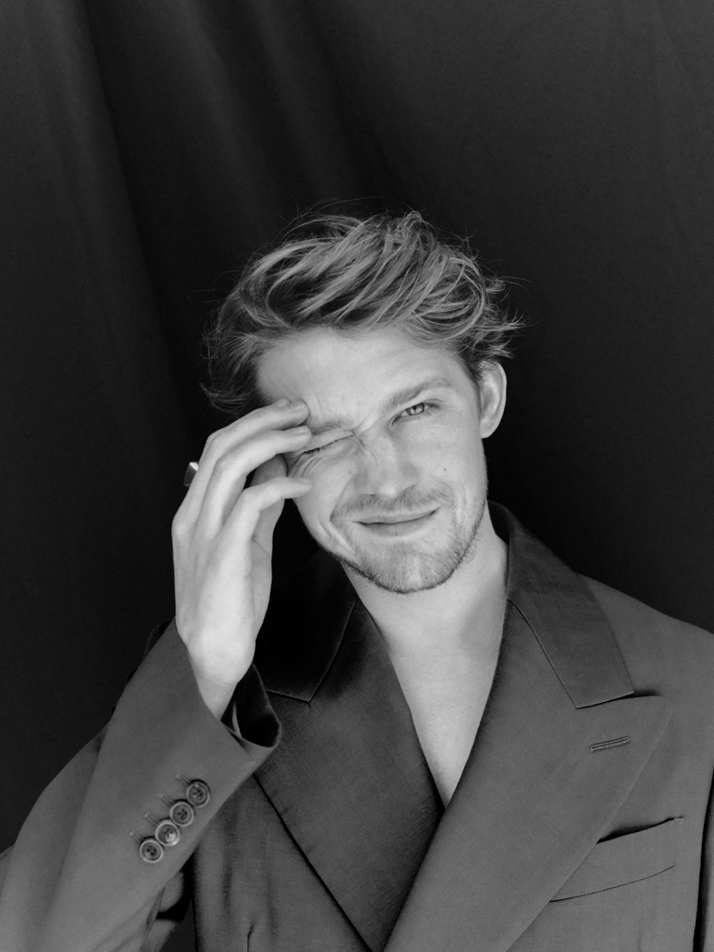 Monochrome portrait of a young adult male in an oversized blazer, winking with blond hair against a dark background in a studio setting.