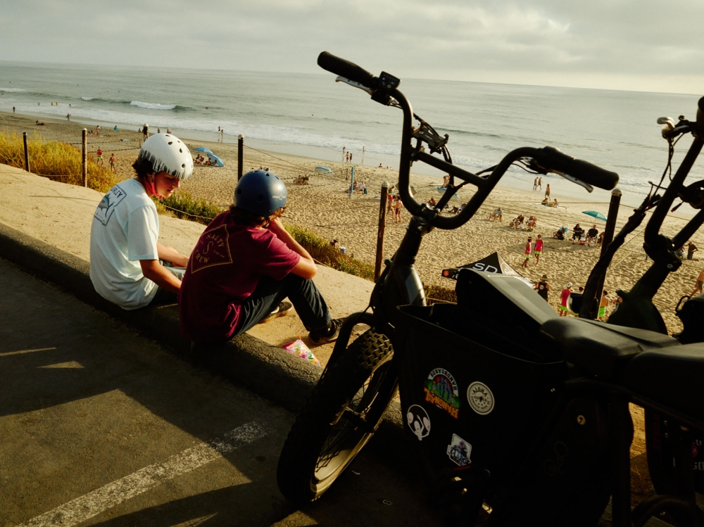 Two teenagers with helmets sitting near electric bikes by beach. Ocean waves and people on sand in background on a sunny day.