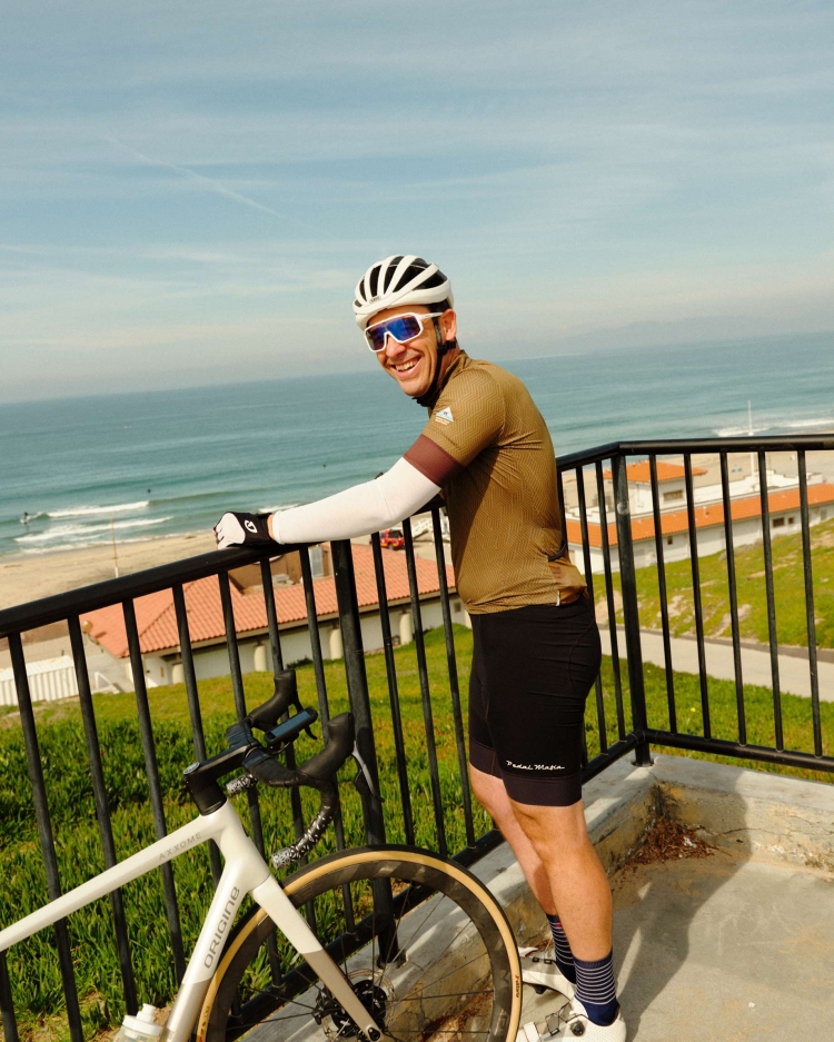 Smiling cyclist in cycling gear poses on a balcony with a bicycle, overlooking a beach, ocean, and blue sky on a sunny day.