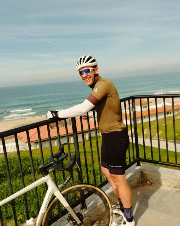 Smiling cyclist in cycling gear poses on a balcony with a bicycle, overlooking a beach, ocean, and blue sky on a sunny day.