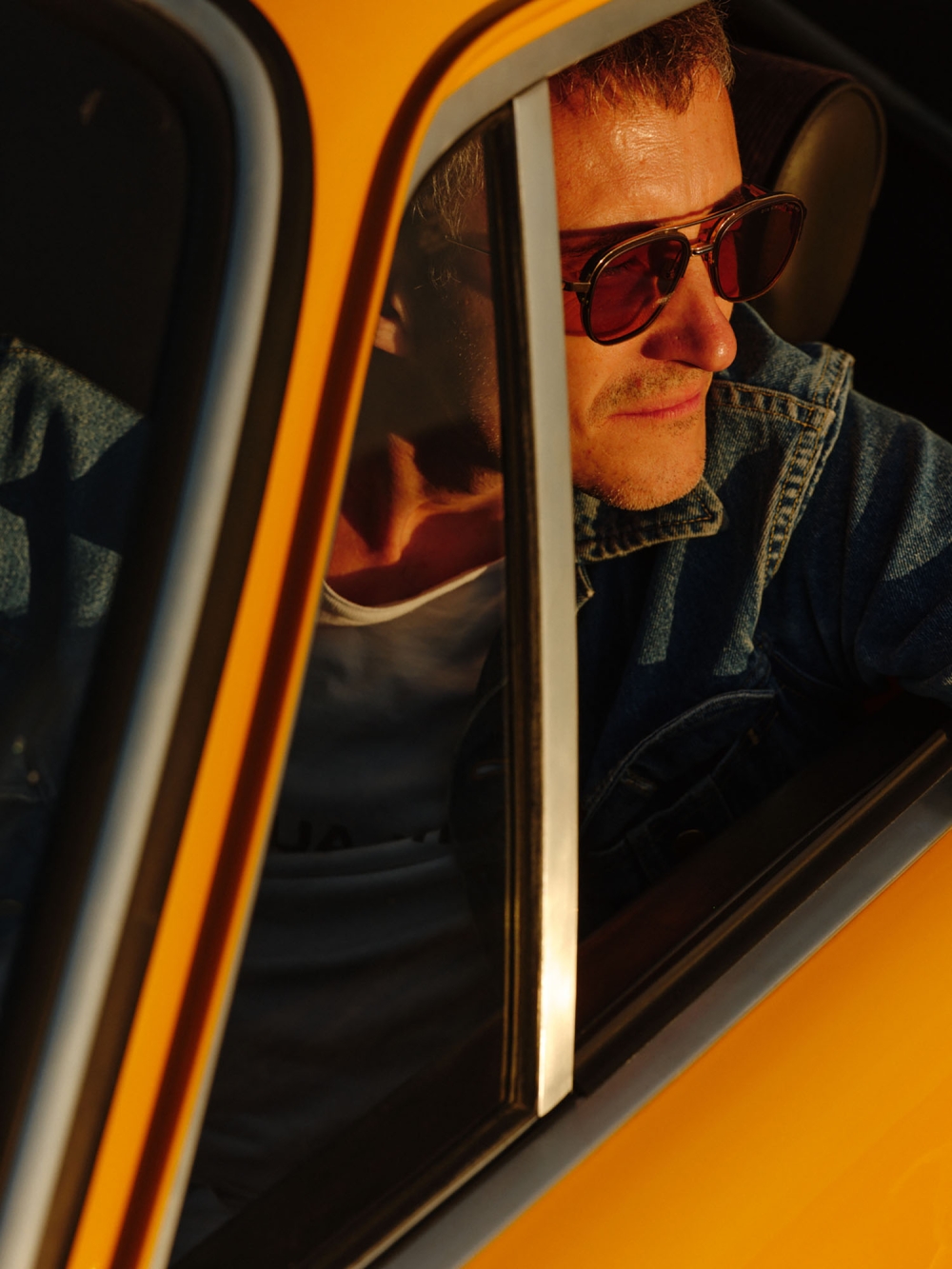 Profile view of a man with sunglasses in a vintage car, captured with natural sunlight and a shallow depth of field, highlighting 1970s style.