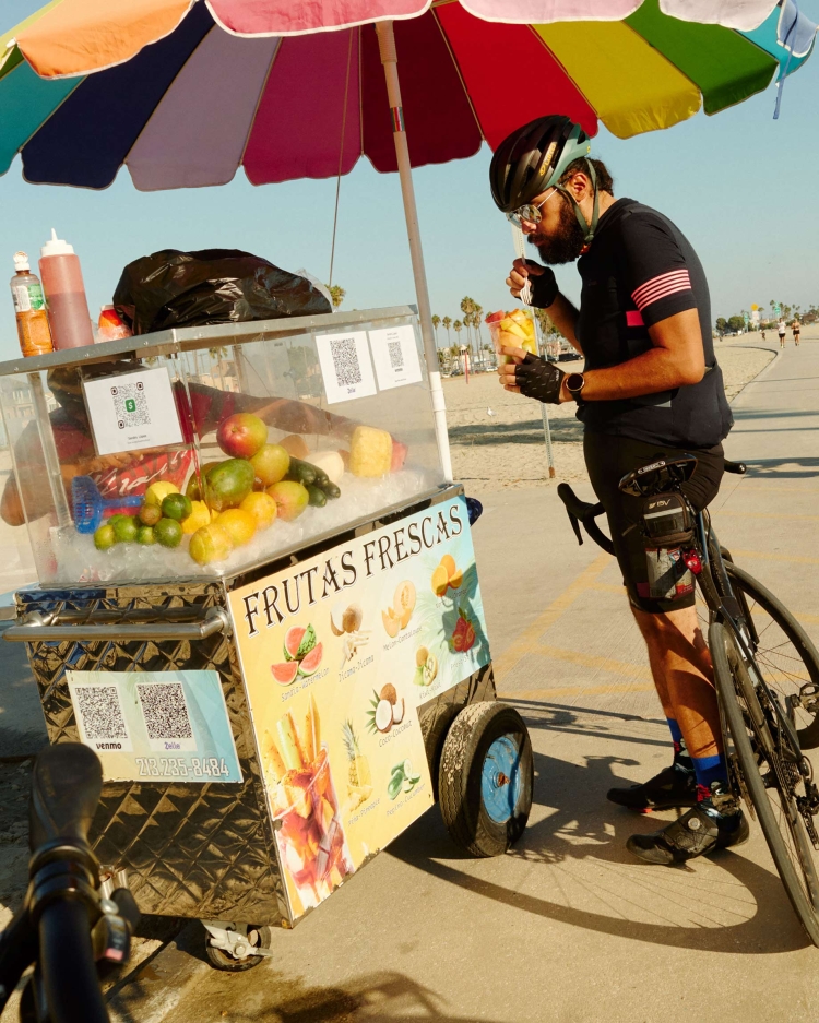 Bearded cyclist in helmet enjoys fruit cup from stand under colorful umbrella by beach path on sunny day, bicycle parked nearby.