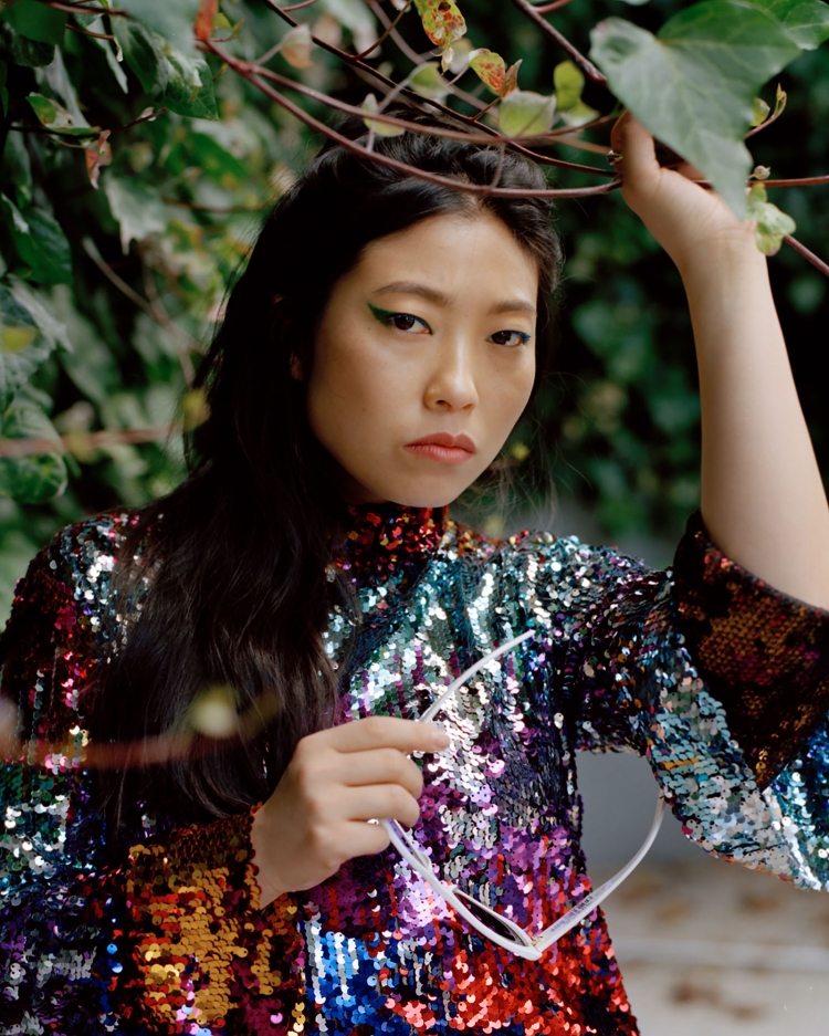 Portrait of a young Asian woman with dark hair, wearing a colorful sequined top, holding white glasses, with green foliage background.
