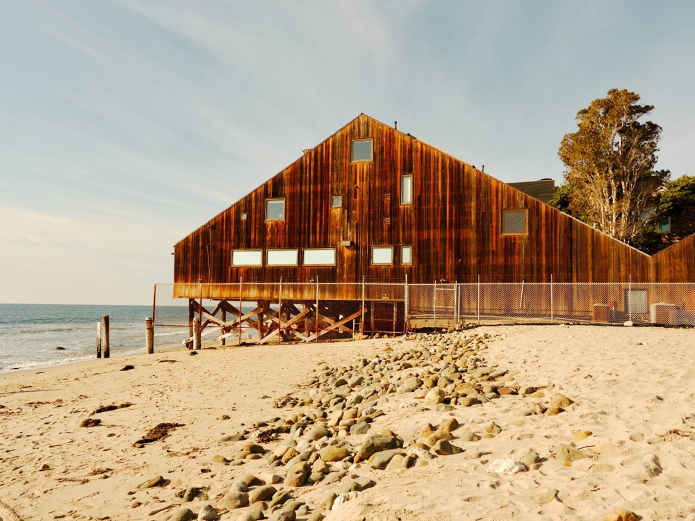 Exterior of wooden beach house on stilts near the ocean with a stone path, taken on a sunny day with a clear sky above.