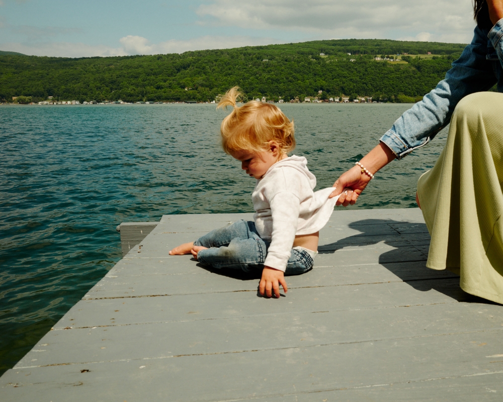 Blond toddler sits on a wooden pier by a lake, holding onto the shirt of a woman in a light-green skirt; forested hills in background.