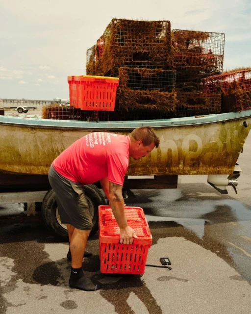 Adult male in a red shirt moving orange baskets from a boat onto a paved surface with other crab traps visible in the background.