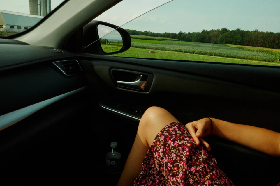 Woman relaxes in car, wearing a floral dress. View of farmland and fields through the car window on a sunny day.