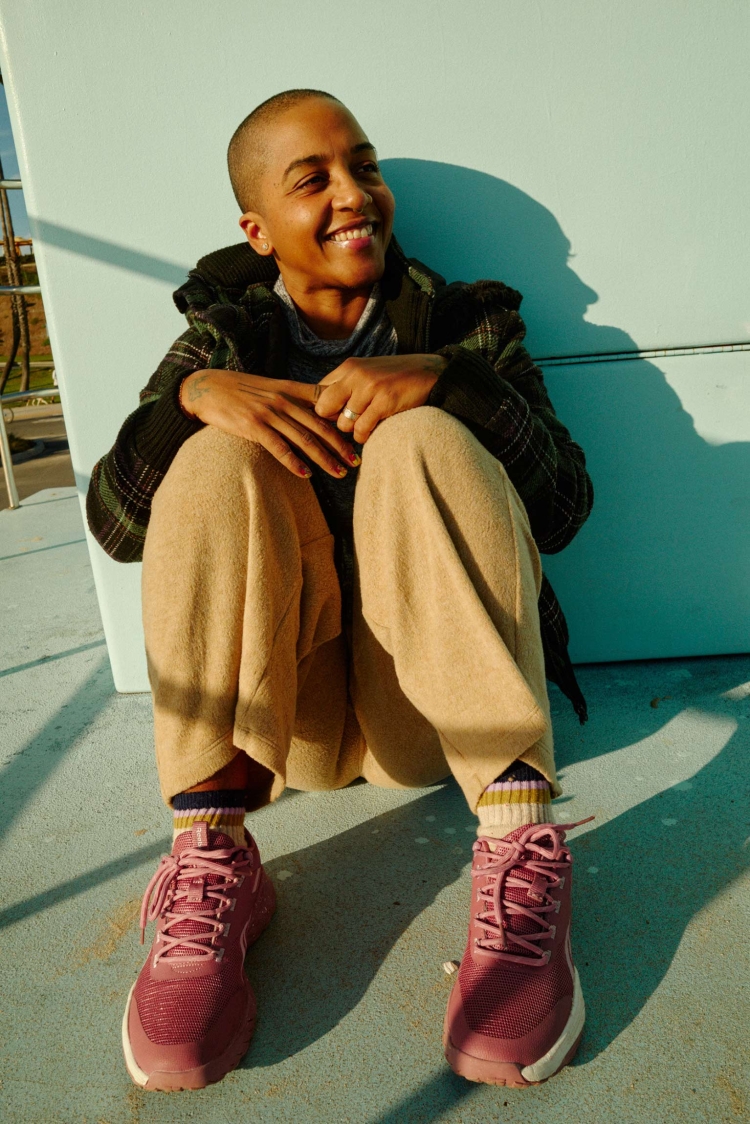 Woman with short hair sits smiling in an outdoor setting, wearing a patterned jacket, tan pants, socks and pink athletic shoes.