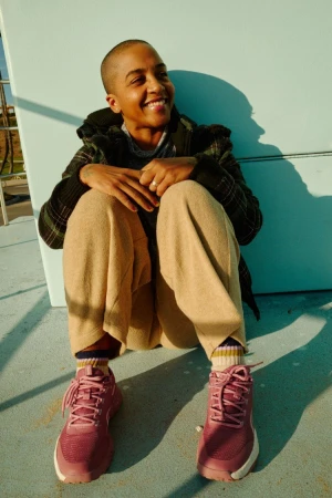 Smiling person with a shaved head wearing a dark patterned jacket, beige trousers, and pink sneakers, seated outdoors in sunlight.