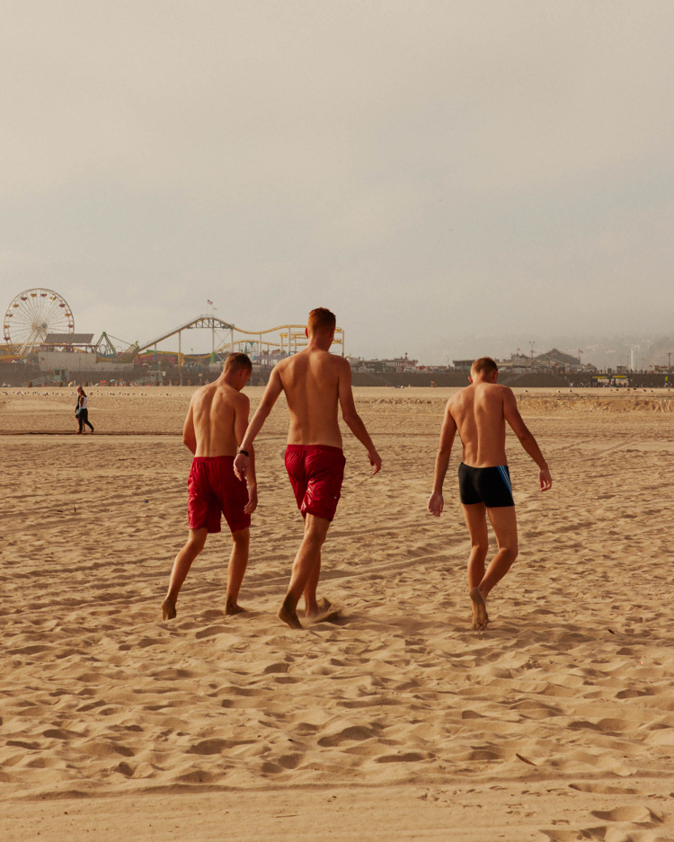 Three adult males walk on a sandy beach with a distant amusement park featuring a Ferris wheel during a sunny day.